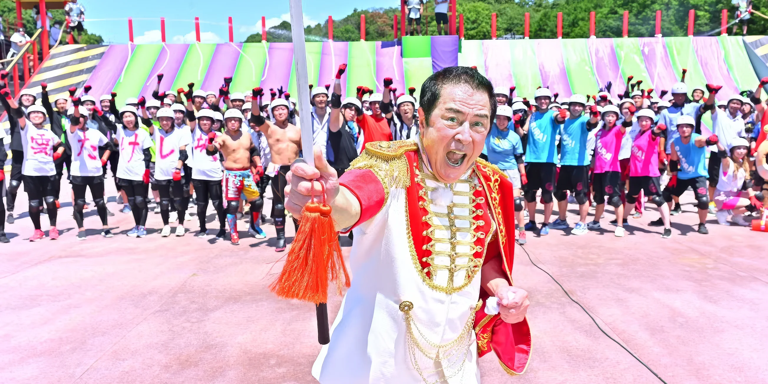 General Tani stands in front of a group of contestants, with the Slippery Wall in the background, on the set of the new Takeshi's Castle 2023 series